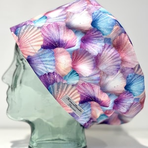 May include: A scrub cap featuring a repeating pattern of seashells in shades of purple, pink, and blue. The fabric has a subtle glittery appearance throughout the shell design. A small rectangular tag with the text Howellmade Devon is attached.