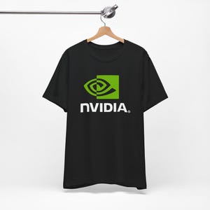 May include: Black t-shirt featuring the NVIDIA logo. The logo includes a green square with a stylized eye design and the word "NVIDIA" in white, sans-serif font. The shirt is hanging on a wooden hanger.