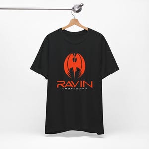 May include: Black t-shirt with a red graphic of a stylized bird or bat and the word "RAVIN" above "CROSSBOWS" in red. The shirt is hanging on a wooden hanger.