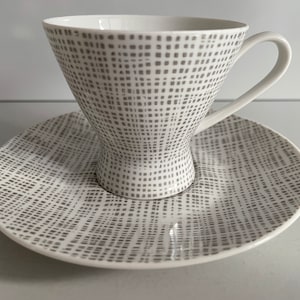 Puede incluir: Juego de taza y platillo de té de cerámica blanca. La taza tiene una forma acampanada y un asa. Ambas piezas están decoradas con un patrón de cuadrados grises. El platillo es redondo y complementa el diseño de la taza. Una adición elegante a cualquier cocina.