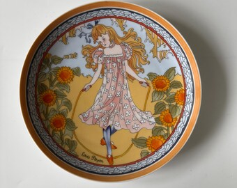 May include: Decorative plate featuring a young girl skipping rope amidst sunflowers. The girl wears a pink dress with white floral accents. The plate has an orange and blue border, with the artist's signature at the bottom.