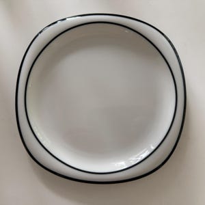 May include: A white ceramic plate with a black rim. The plate has a simple, elegant design, suitable for serving meals. The plate is round and has a slightly raised edge. The black rim provides contrast to the white surface.