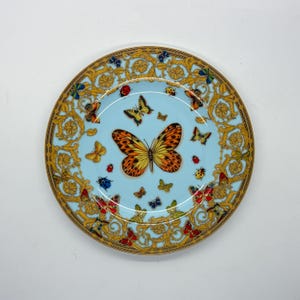 Puede incluir: Un plato decorativo redondo con un centro azul claro y un borde dorado. El plato presenta una gran mariposa naranja y amarilla en el centro, rodeada de mariposas y mariquitas más pequeñas. El borde tiene un diseño dorado ornamentado.