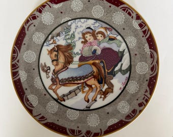 May include: Decorative plate featuring a winter scene with a horse-drawn sleigh carrying two children. The plate has a gray border with white snowflake designs and a maroon rim. The horse is brown, and the sleigh is burgundy.
