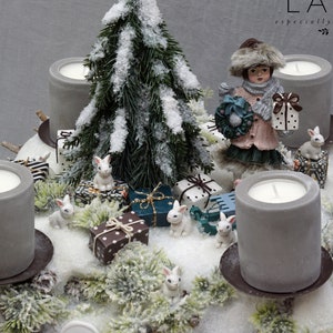 Könnte beinhalten: Ein grauer Holzsockel mit einer schneeweißen Oberfläche, verziert mit einem kleinen schneebedeckten Tannenbaum, einer Porzellanfigur eines Mädchens, das einen Kranz und ein Geschenk hält, und vier grauen Kerzen. Außerdem sind kleine weiße Kaninchen und verpackte Geschenke um den Sockel herum verstreut. Der Sockel ist mit einem weißen Etikett mit dem Text "LAVI especially for me" verziert.