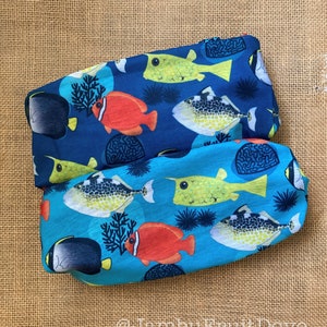 Op de afbeelding: Twee stoffen haarbanden met een levendig aquatisch ontwerp. De haarbanden hebben kleurrijke vissen, koraal en zee-egels op een blauwe en turquoise achtergrond. Het ontwerp bevat gele, rode, zwarte en witte vissen. De tekst "@JambuFruitDove" is zichtbaar.
