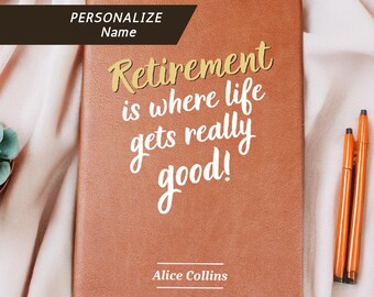 May include: A brown leather-bound journal with the text "Retirement is where life gets really good!" in gold script. The name "Alice Collins" is printed below the quote. Three orange pens are next to the journal. A succulent plant is in the background.