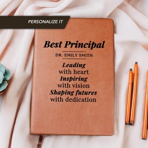 Pode incluir: Um caderno marrom com a inscrição "Best Principal" e "Leading with heart, Inspiring with vision, Shaping futures with dedication." Várias canetas laranja estão ao lado do caderno. Uma planta suculenta está ao fundo.