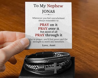 Puede incluir: Una pulsera de cuero negro con un dije de cruz plateada, presentada en una caja de regalo negra. Una tarjeta en el fondo dice "To My Nephew Jonas" con un mensaje inspirador sobre la oración.