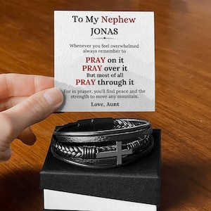 Könnte beinhalten: Ein schwarzes Lederarmband mit einem silbernen Kreuzanhänger, präsentiert in einer schwarzen Geschenkbox. Eine Karte im Hintergrund trägt die Aufschrift "To My Nephew Jonas" mit einer inspirierenden Botschaft über das Gebet.