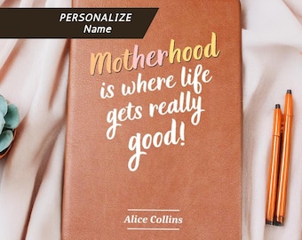 May include: A brown journal with the text "Motherhood is where life gets really good!" in white and yellow script. The journal has a ribbon bookmark and is accompanied by orange pens. The name "Alice Collins" is printed at the bottom.