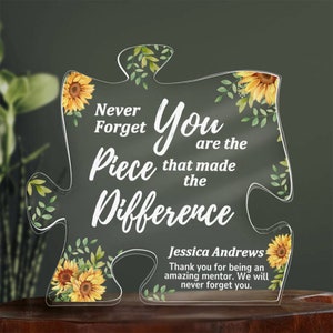 Puede incluir: Una pieza de rompecabezas de acrílico transparente con un diseño de girasol y el texto "Never Forget You are the Piece that made the Difference". También se incluye el texto "Jessica Andrews Thank you for being an amazing mentor. We will never forget you."