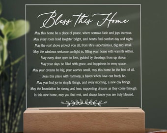 May include: A white acrylic sign with a wooden base, featuring a poem about home and blessings. The sign reads "Bless this Home" and includes a verse about finding peace, joy, and comfort in a new home.