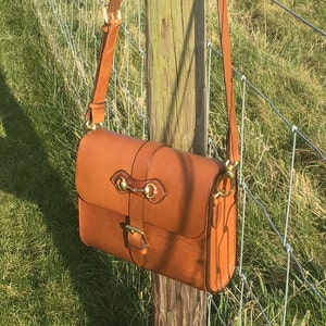 May include: A brown leather satchel bag with a brass buckle and hardware. The bag has a long strap and is hanging on a wooden fence post.