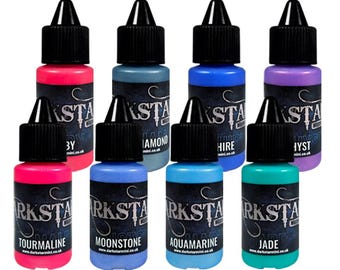May include: A collection of eight small, colorful bottles with black caps. Each bottle is labeled with a different color name, including Tourmaline, Moonstone, Aquamarine, and Jade. The bottles are arranged in a visually appealing display.
