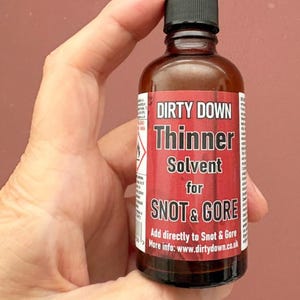 May include: A brown glass bottle with a black cap, held in a hand. The label reads "DIRTY DOWN Thinner Solvent for SNOT & GORE." Additional text includes instructions and a website address.