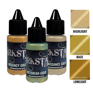 Puede incluir: Tres pequeños botes de pintura con tapas negras y etiquetas, que muestran tonos dorados. Las etiquetas dicen "Regency Gold", "Victorian Gold" y "Renaissance Gold". También se muestran muestras de color para resaltado, base y sombra.