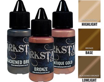 May include: Three bottles of Darkstar Miniatures metallic paints: Blackened Bronze, Bronze, and Antique Gold. Each bottle has a black cap and a label with the brand name and color. Color swatches for Highlight, Base, and Lowlight are also shown.