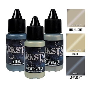 Puede incluir: Tres pequeños botes de pintura con tapas negras y etiquetas. Los botes están etiquetados como "Steel", "Silver Verde" y "Old Silver". También se muestran muestras de color para "Highlight", "Base" y "Lowlight". El texto "DARKSTAR" y "www.darkstarmini.co.uk" son visibles.