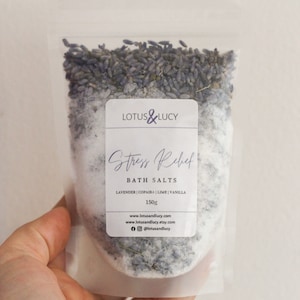May include: A clear plastic pouch filled with white bath salts and lavender buds. The pouch has a white label with the text "LOTUS&LUCY", "Stress Relief", "BATH SALTS", "LAVENDER | COPAIBA | LIME | VANILLA", "150g", "www.lotusandlucy.com", "www.lotusandlucy.etsy.com", and "@lotusandlucy".