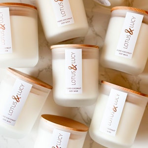 May include: Overhead view of several white candles with wooden lids. Each candle has a label that reads "LOTUS & LUCY PURE COCONUT SOY WAX." The candles are in clear glass jars, set against a white marble surface.