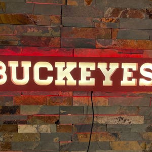 Puede incluir: Un letrero rectangular rojo con la palabra "BUCKEYES" en letras blancas grandes e iluminadas. El letrero está montado en una pared de piedra texturizada y multicolor. Un cable de alimentación negro sale de la parte inferior.