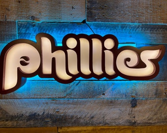 May include: A white illuminated sign with the word "Phillies" in a retro font, mounted on a rustic wooden wall with blue lighting.