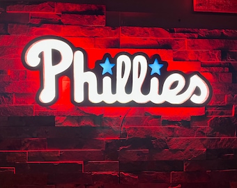 May include: A white illuminated sign with the word "Phillies" in a cursive font. Two blue stars are above the word "Phillies". The sign is mounted on a red brick wall.