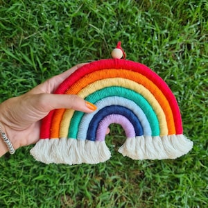 May include: A colorful rainbow wall hanging made of yarn with a wooden bead at the top. The rainbow is made of red, orange, yellow, green, blue, indigo, and violet yarn. The rainbow has white fringe at the bottom.