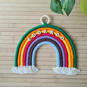 May include: A colorful rainbow wall hanging made of yarn with the name "EDOARDO" spelled out in wooden letters. The rainbow has a white fringe at the bottom and is hung from a wooden ring.