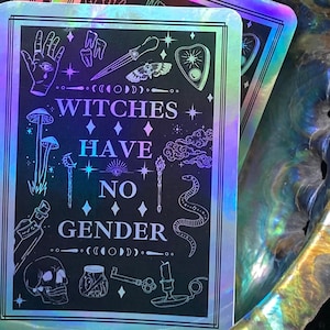 May include: A holographic sticker with a black background and white text that reads "Witches Have No Gender". The sticker is decorated with various witchy symbols, including a skull, a snake, a key, a candle, a crescent moon, a hand with an eye, a moth, a cauldron, and a mushroom.