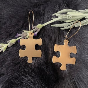 May include: A pair of gold-toned puzzle piece shaped earrings. The earrings are hanging from a fuzzy black surface.
