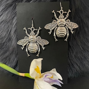 May include: A pair of silver bee earrings with detailed wings and bodies. The earrings are displayed on a black background.