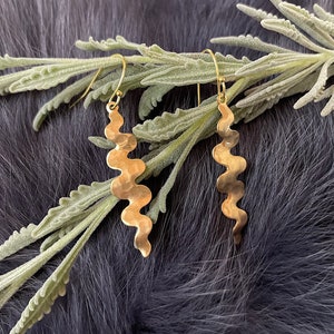 以下が含まれることがあります： 金色の波型のイヤリングのペア。ハンマーで叩いたような質感です。イヤリングはシンプルなフックから吊り下げられ、緑の葉の上に、暗く質感のある背景に展示されています。イヤリングはユニークで有機的な形をしています。