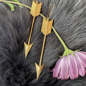 May include: A pair of gold arrow-shaped earrings with a feather design. The earrings are hanging from a fuzzy gray surface.