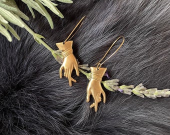 May include: A pair of gold-colored hand-shaped earrings. Each earring features a detailed hand design with a wrapped wrist and a long, thin hook. The earrings are displayed on a dark, textured surface with sprigs of greenery.