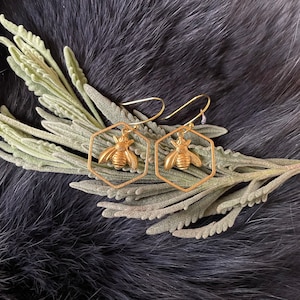 May include: A pair of gold-toned earrings featuring a bee design inside a hexagonal frame. The earrings are hanging from a silver hook.