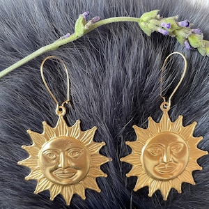 May include: A pair of gold-colored sun face earrings with a detailed design. The earrings have a hook closure and are displayed on a dark, textured background with a sprig of lavender.