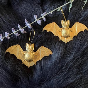 May include: A pair of gold-colored bat earrings. The bats have detailed wings and bodies.