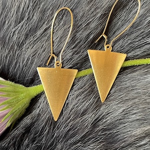 May include: A pair of gold triangle earrings with a simple design. The earrings are hanging from a fuzzy surface.