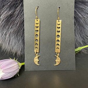 May include: Gold tone earrings with a crescent moon design. The earrings feature a long, rectangular bar with a series of crescent moon cutouts. The bottom of the bar is attached to a crescent moon charm.