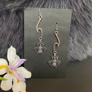 May include: A pair of silver earrings with a bee design. The earrings are hanging from a silver hook and have a wave-like design.