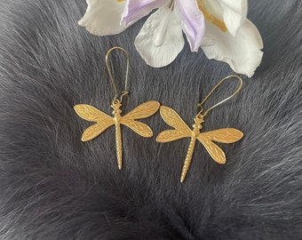 May include: A pair of gold-toned dragonfly earrings with delicate wings and a dangling design. The earrings are shown against a gray furry background.