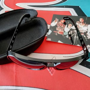 May include: Black sunglasses with red lenses and a black case. The sunglasses have a sleek, modern design with a curved frame and a small tag that says UV 400. The case is open, revealing a soft interior.