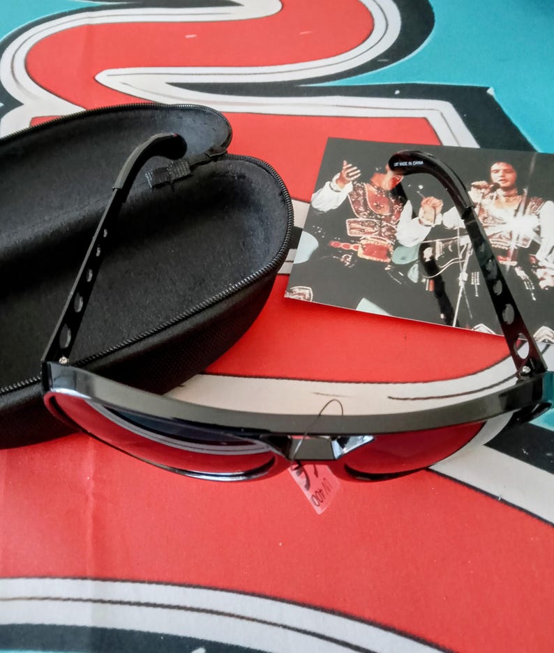 May include: Black sunglasses with red lenses and a black case. The sunglasses have a sleek, modern design with a curved frame and a small tag that says UV 400. The case is open, revealing a soft interior.