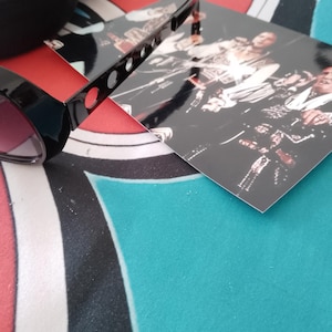May include: Black sunglasses with pink lenses and a black case are placed on a patterned surface. A black, rectangular object with holes is also visible, along with a photograph of a band.