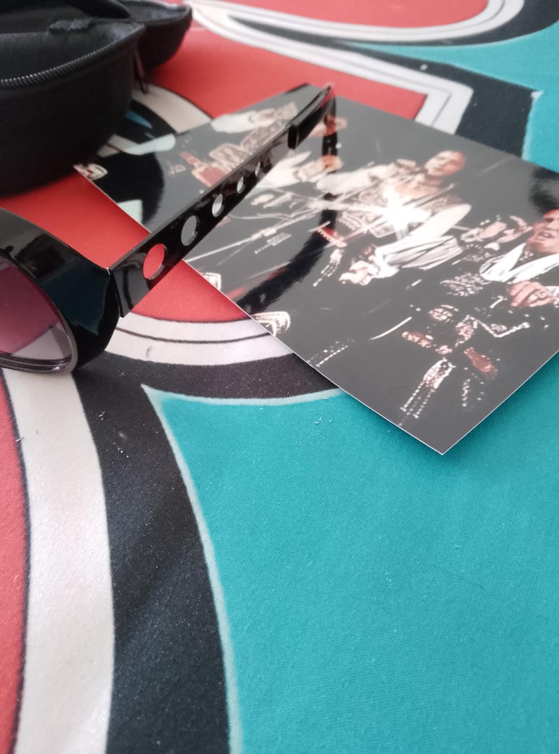May include: Black sunglasses with pink lenses and a black case are placed on a patterned surface. A black, rectangular object with holes is also visible, along with a photograph of a band.