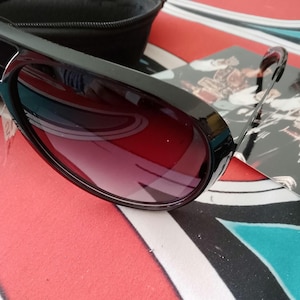 May include: Black sunglasses with a dark red gradient lens. The sunglasses have a thick, rectangular frame and are resting on a colorful patterned surface. A black case is visible in the background.