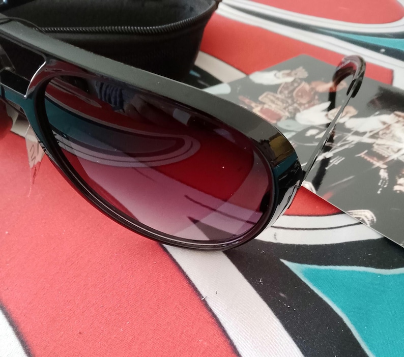 May include: Black sunglasses with a dark red gradient lens. The sunglasses have a thick, rectangular frame and are resting on a colorful patterned surface. A black case is visible in the background.