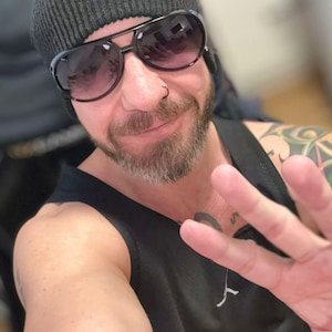 May include: A person wearing a black beanie and sunglasses with a dark frame. The person is also wearing a black tank top. The person is holding up their hand with three fingers extended.